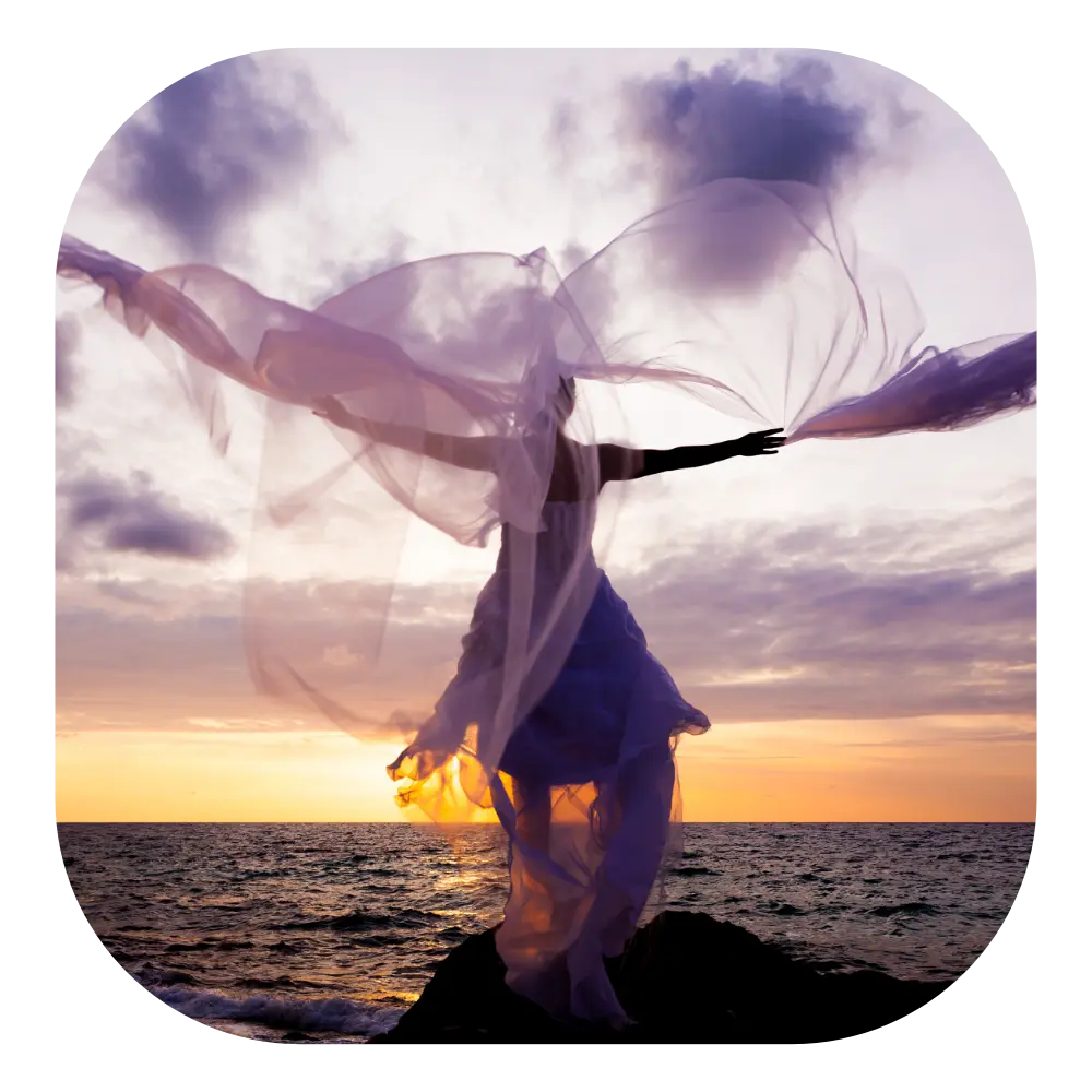 A flowing, translucent white and lavender fabric drapes dynamically around a figure standing on a rocky shore at sunset.